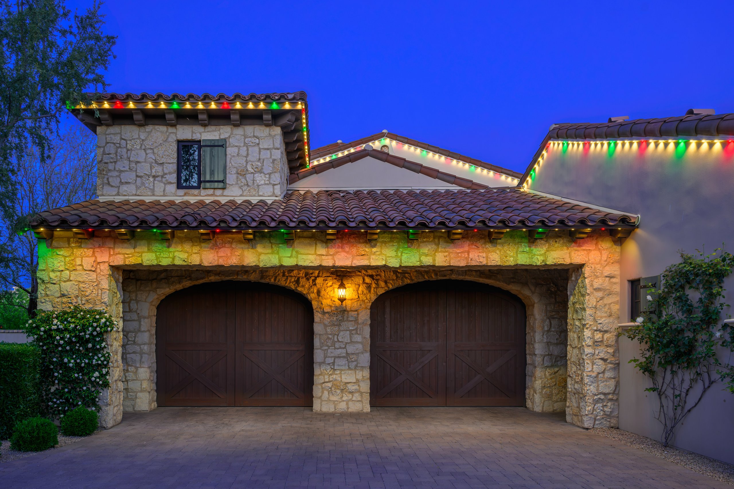 Red and green permanent trim holiday lighting on a stone two-car garage at dusk in Phoenix